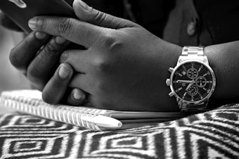 Close-up of hands holding a phone over a notebook. A wristwatch is visible on one of the hands, and the background includes a patterned fabric.