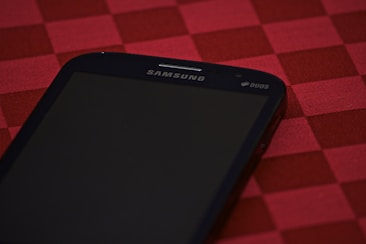 A smartphone with the brand name Samsung and Duos written on the top side, placed on a red checkered tablecloth. The phone's screen is dark and not illuminated.