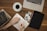 A person is writing in a planner or diary on a wooden table surrounded by various items. These include a cup of black coffee, a pair of eyeglasses, a stack of books, a laptop, a tablet, and several printed photographs. The scene suggests a cozy, organized workspace.