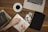 A person is writing in a planner or diary on a wooden table surrounded by various items. These include a cup of black coffee, a pair of eyeglasses, a stack of books, a laptop, a tablet, and several printed photographs. The scene suggests a cozy, organized workspace.