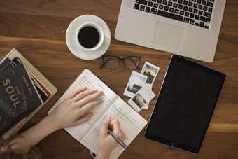 A person is writing in a planner or diary on a wooden table surrounded by various items. These include a cup of black coffee, a pair of eyeglasses, a stack of books, a laptop, a tablet, and several printed photographs. The scene suggests a cozy, organized workspace.