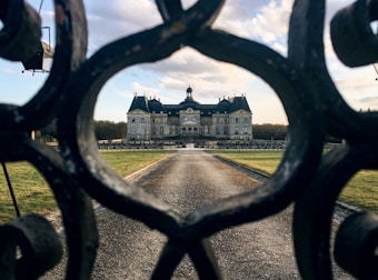 A grand chateau is framed by ornate, wrought iron gates. The building sits at the end of a long, straight pathway surrounded by manicured lawns. The sky is partly cloudy with a gentle light illuminating the scene.