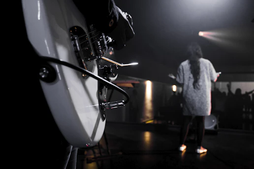 A close-up of Aszait playing guitar on stage, bathed in soft white light with a hint of rebellious red in the background.