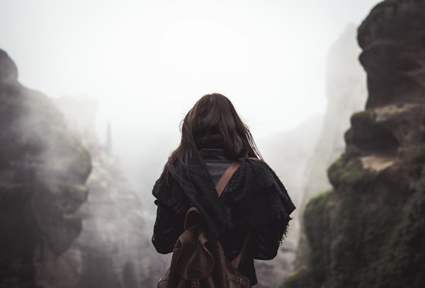 A woman with a brown backpack standing with her back to the camera, looking out at mountains