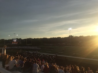 A football field under a clear sky during sunset, with bright stadium lights illuminating the scene. A crowd of spectators is seated in the foreground, watching an event on the field, possibly a marching band performance, with a scoreboard visible on the left side.