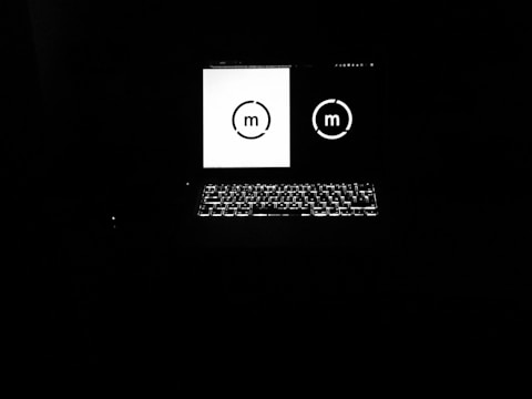 A laptop is open in a dark environment, with the screen displaying two similar circular logos, one in black on a white background and the other in white on a black background. The keyboard is backlit, providing a subtle glow.
