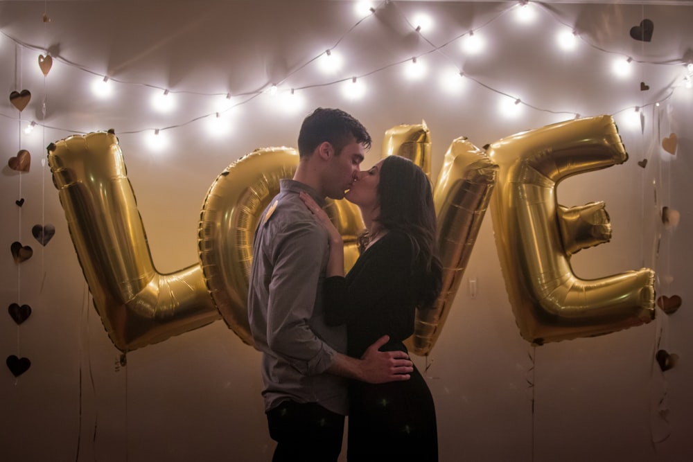 Un couple s’embrassant devant des ballons de couleur or qui épelent « Amour ».