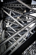Intricate steel beams and trusses form a complex lattice structure, showcasing industrial design and engineering. The interlocking metal components create geometric patterns with rivets visible throughout the structure.