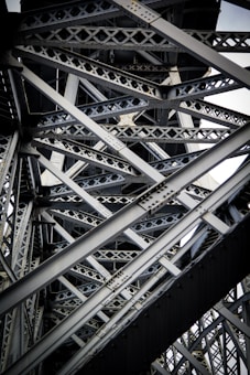 Intricate steel beams and trusses form a complex lattice structure, showcasing industrial design and engineering. The interlocking metal components create geometric patterns with rivets visible throughout the structure.