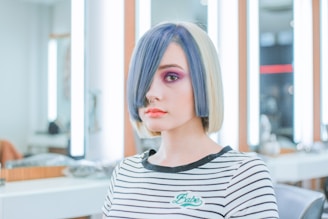 A young person with a striking hairstyle featuring a bob cut, with half blue and half blonde hair. They have bold makeup with bright pink eyeshadow and orange lipstick, and they are wearing a striped shirt. The background appears to be a modern salon with bright lights and mirrors.