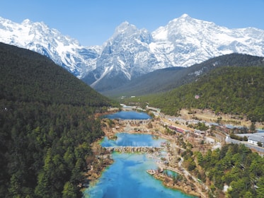A picturesque valley with a series of blue lakes interspersed with greenery and surrounded by majestic snow-capped mountains. The landscape is lush, with dense forests on one side and a few structures visible.