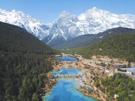 A picturesque valley with a series of blue lakes interspersed with greenery and surrounded by majestic snow-capped mountains. The landscape is lush, with dense forests on one side and a few structures visible.