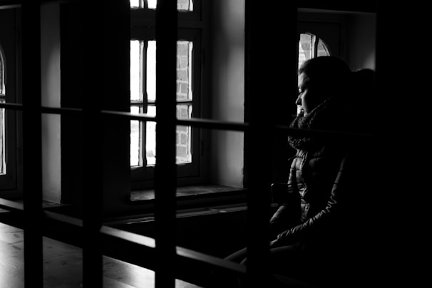 A person sits in a dimly lit room, gazing out of a window. Shadows and light create a strong contrast, highlighting the contemplative expression on the person's face. The scene gives a feeling of solitude and introspection.