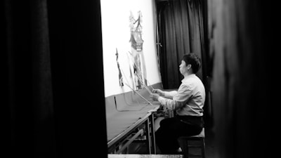 A person is sitting on a stool in a dimly lit room, operating shadow puppets behind a backlit screen. The setting suggests a traditional shadow puppet performance. The individual is focused and appears to be manipulating the puppets using rods.