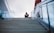 A solitary person is seated on a flight of wide concrete stairs that leads up to a modern building. The individual is wearing a cap and casual clothing, with their head slightly bowed and arms resting on their knees in a contemplative posture. The staircase is flanked by glass railings, and the sky above is overcast, creating a subdued atmosphere.