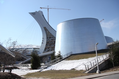 A modern architectural structure featuring a curved, white tower with cables resembling a suspension bridge. Next to it, a large cylindrical building with shiny, metallic walls. There is a construction crane in the background, and patches of snow on the ground surrounded by sparse vegetation.