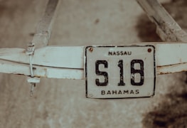 A weathered license plate with the inscription 'NASSAU S16 BAHAMAS' is attached to a worn, white metal structure. The background appears to be a textured or sandy surface.