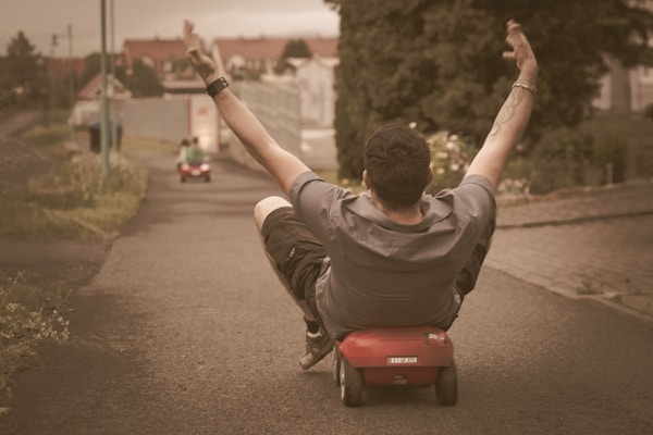 A person is riding a small toy car down a paved path with arms raised in excitement. The scene appears playful and carefree, taking place outdoors in what looks like a residential area.