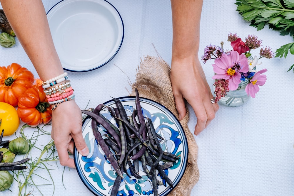 pessoa segurando prato de cerâmica floral branca e azul com feijão de corda no topo perto de tomates de herança