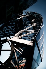 A close-up view of a modern architectural structure featuring a complex steel framework and curved glass elements. The design includes intersecting metal beams and an array of reflective surfaces, creating a dynamic and futuristic appearance.