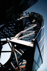 A close-up view of a modern architectural structure featuring a complex steel framework and curved glass elements. The design includes intersecting metal beams and an array of reflective surfaces, creating a dynamic and futuristic appearance.