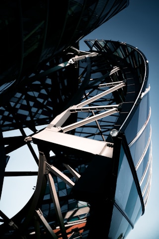 A close-up view of a modern architectural structure featuring a complex steel framework and curved glass elements. The design includes intersecting metal beams and an array of reflective surfaces, creating a dynamic and futuristic appearance.