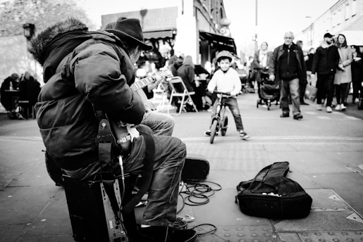A street musician playing an electric guitar while sitting on a small amplifier. A young child on a bicycle approaches, surrounded by a busy street scene with people walking by and a cafe in the background. Musical cables extend from the guitar, with an open guitar case on the ground potentially for tips.
