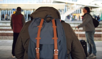 A person with a large blue backpack stands on a train platform, facing the tracks. There are two other people visible, one on the left wearing a red vest and another on the right with a suitcase, suggesting they are also travelers. The background features a train station with several people and a large, modern architectural structure.