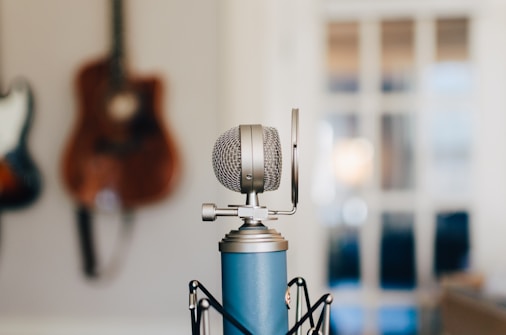 A close-up of a blue studio microphone in focus, with a pop filter attached. In the background, a blurred guitar hangs on the wall, adding an artistic and musical ambiance.