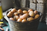 A rustic metal bucket filled with brown eggs is placed on a table. Some eggs have specks of dirt and straw attached, suggesting they are freshly gathered. The background displays blurred stacks of cardboard egg trays, emphasizing a farm-like environment.