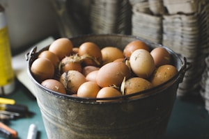 A rustic metal bucket filled with brown eggs is placed on a table. Some eggs have specks of dirt and straw attached, suggesting they are freshly gathered. The background displays blurred stacks of cardboard egg trays, emphasizing a farm-like environment.