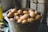 A rustic metal bucket filled with brown eggs is placed on a table. Some eggs have specks of dirt and straw attached, suggesting they are freshly gathered. The background displays blurred stacks of cardboard egg trays, emphasizing a farm-like environment.