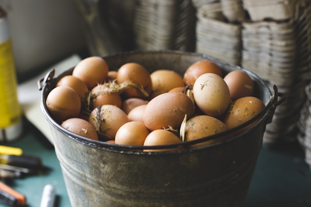 A rustic metal bucket filled with brown eggs is placed on a table. Some eggs have specks of dirt and straw attached, suggesting they are freshly gathered. The background displays blurred stacks of cardboard egg trays, emphasizing a farm-like environment.