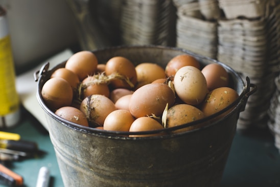 A rustic metal bucket filled with brown eggs is placed on a table. Some eggs have specks of dirt and straw attached, suggesting they are freshly gathered. The background displays blurred stacks of cardboard egg trays, emphasizing a farm-like environment.