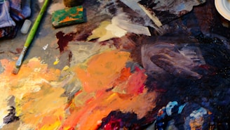 A vibrant artist's palette with an assortment of mixed acrylic paints in various shades of orange, yellow, brown, and blue. There is a well-used paintbrush with a green handle laid across the palette, alongside other painting tools and supplies. The paint is thickly applied, creating a textured surface with swirls and strokes indicating artistic work in progress.