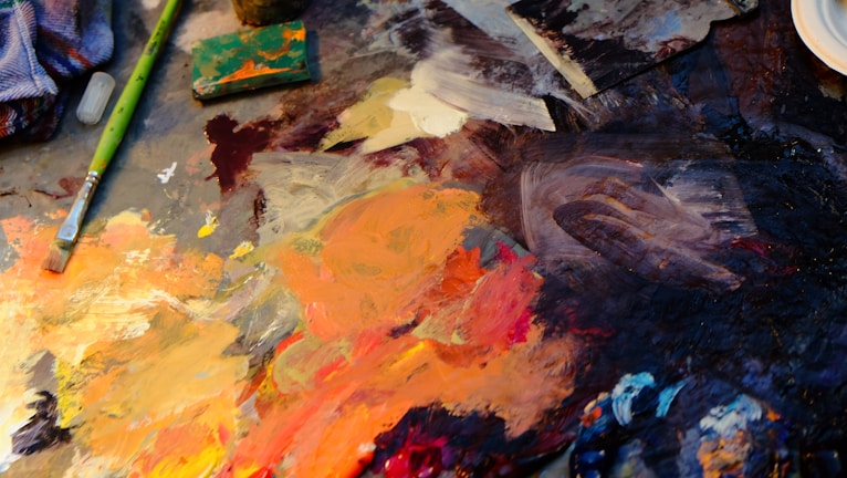 A vibrant artist's palette with an assortment of mixed acrylic paints in various shades of orange, yellow, brown, and blue. There is a well-used paintbrush with a green handle laid across the palette, alongside other painting tools and supplies. The paint is thickly applied, creating a textured surface with swirls and strokes indicating artistic work in progress.