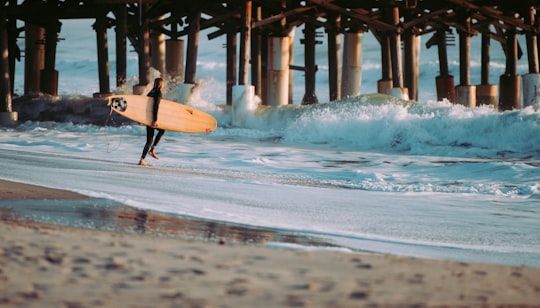 person holding surfboard standing on seashore in front of wave near wooden dock in Cocoa Beach United States