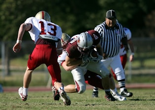 Two football players are in action on the field, with one wearing a maroon jersey and helmet tackling another in a white jersey with the number 13. A referee in a striped uniform stands in the background, and another player is partially visible.