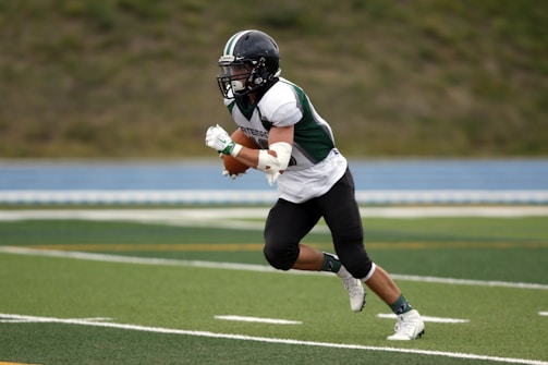 A football player in a dark helmet and green and white uniform is running on a green field, holding a brown football. The player is wearing white cleats and is captured mid-action, seemingly in a sprint. The background features a blurred view of a blue track and grassy area.