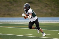 A football player in a dark helmet and green and white uniform is running on a green field, holding a brown football. The player is wearing white cleats and is captured mid-action, seemingly in a sprint. The background features a blurred view of a blue track and grassy area.