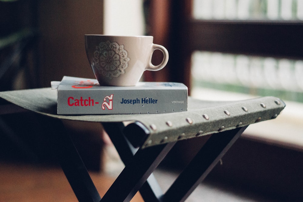 close-up photography of beige ceramic coffee cup on top of Joseph Heller book