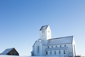 A large white church with a steeply pitched roof and a tall bell tower, set against a clear blue sky. Snow covers the ground and dusts the roof of the building. To the left, there is a smaller wooden building that also has a snow-covered roof.