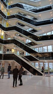 A modern interior with multiple levels connected by zigzagging black staircases. Glass walls create an open and bright atmosphere. Several people are scattered throughout the lobby, some standing in groups, interacting or walking through the space.