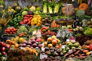 A vibrant market display filled with an assortment of fruits and vegetables, including apples, oranges, asparagus, eggplants, pumpkins, pineapples, and leafy greens. The produce is neatly organized and labeled, showcasing a variety of colors and textures.