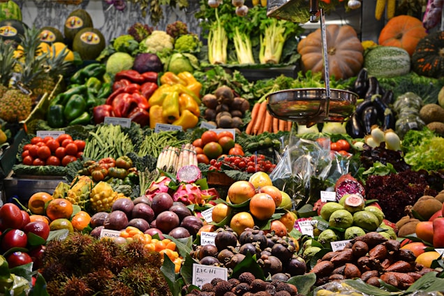 A vibrant market display filled with an assortment of fruits and vegetables, including apples, oranges, asparagus, eggplants, pumpkins, pineapples, and leafy greens. The produce is neatly organized and labeled, showcasing a variety of colors and textures.