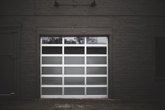 A modern white garage door with several rectangular glass panels is set within a dark brick exterior. The reflection of bare trees is visible in the glass panels, suggesting a winter setting.