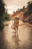 An elderly woman wearing a traditional conical hat and patterned dress walks through a shallow stream. She carries a woven basket and the scene is set against a backdrop of eroded red cliffs with lush greenery. Other figures are visible walking in the distance.