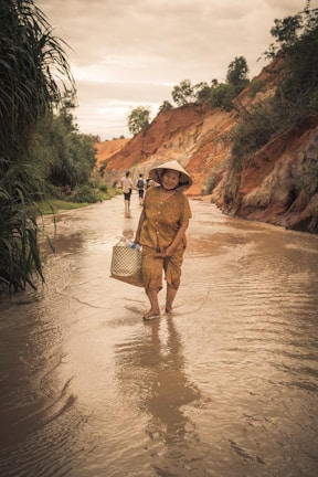 An elderly woman wearing a traditional conical hat and patterned dress walks through a shallow stream. She carries a woven basket and the scene is set against a backdrop of eroded red cliffs with lush greenery. Other figures are visible walking in the distance.