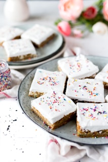 A plate of square-shaped dessert bars with white frosting and colorful sprinkles. The treats are arranged neatly on a gray speckled plate, with a small jar of sprinkles nearby. In the background, there are pink and white flowers, adding a soft, decorative touch.