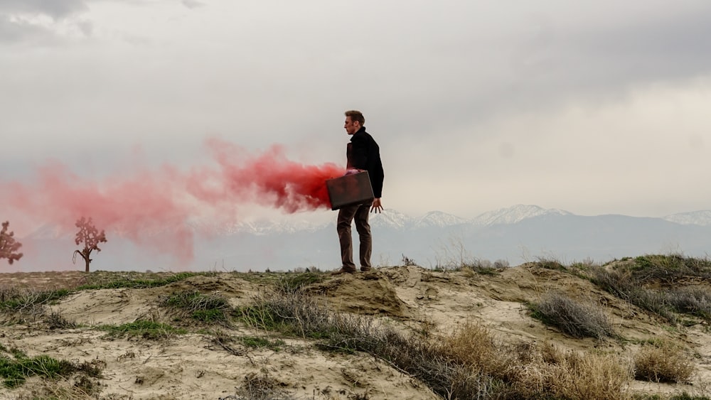 Red smoke escaping a briefcase, held by a business man at the edge of a cliff.